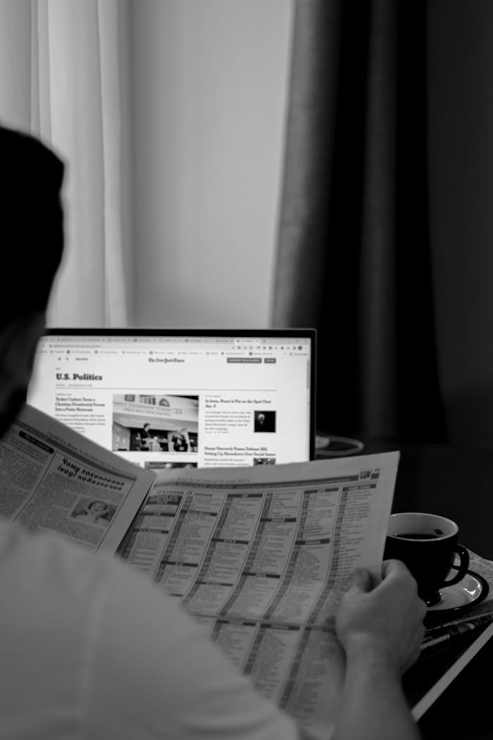 Black and white photo of a man reading a newspaper with a laptop in the background.