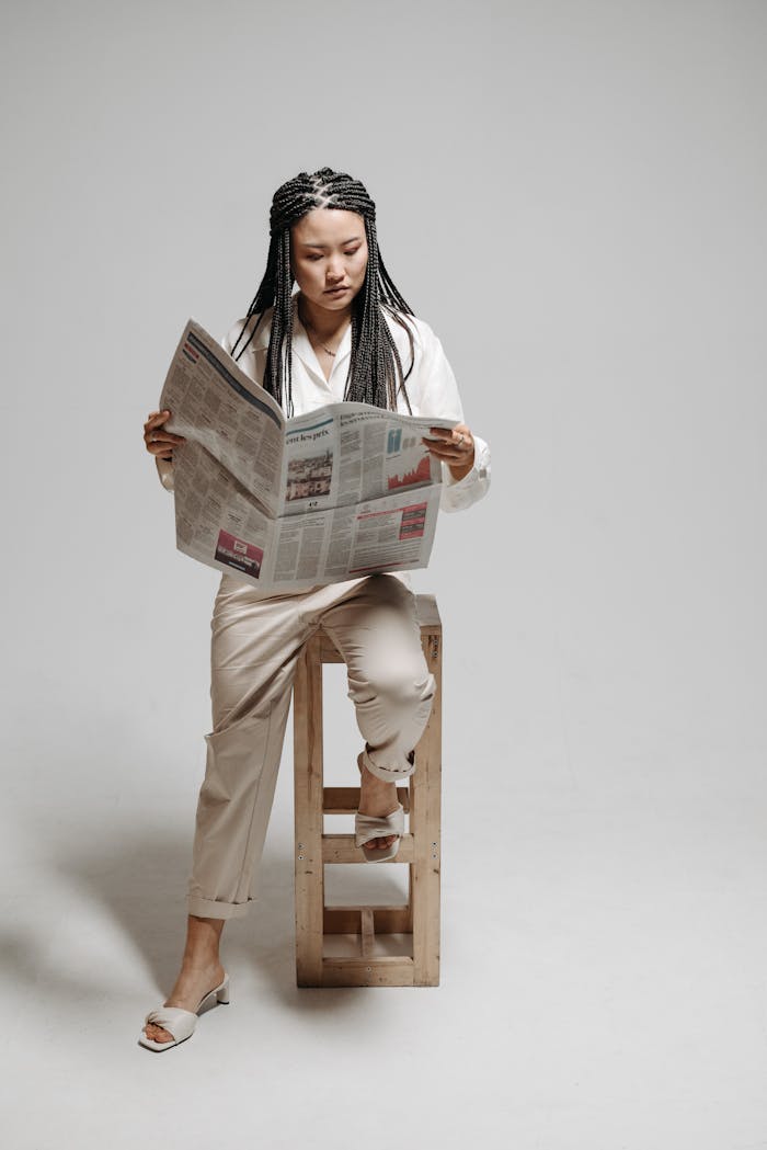An Asian woman with braided hair sits stylishly on a wooden stool reading a newspaper in a minimalist studio setting.